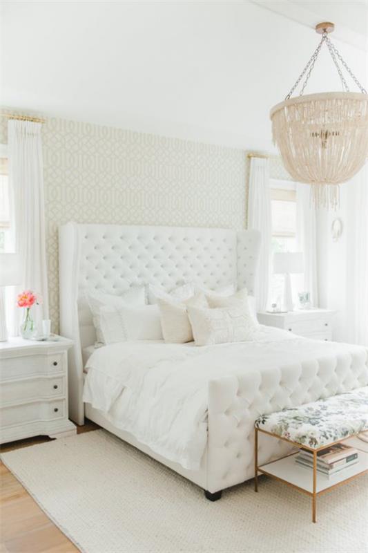 Λαμπερό υπνοδωμάτιο σχεδιασμένο όλο σε λευκό επικαλυμμένο κρεβάτι μαλακό κρεβάτι διακοσμητικά μαξιλάρια πολυέλαιος ροζ λουλούδι σε βάζο από το κομοδίνο