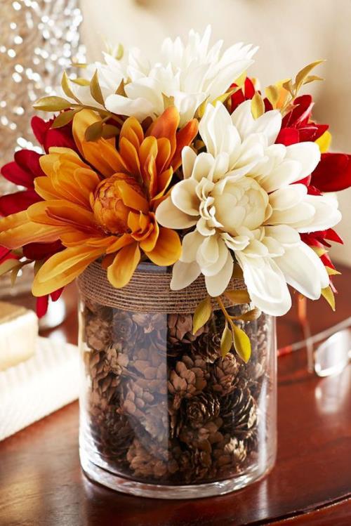 Φθινοπωρινή διακόσμηση με κουκουνάρια μεγάλο ποτήρι ως κεντρικό κομμάτι στο τραπέζι γεμάτο με κουκουνάρια διακοσμημένα με φθινοπωρινά λουλούδια χρυσάνθεμα