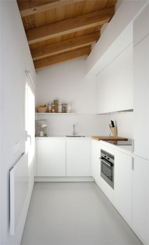 Η κουζίνα L σε λευκό χρώμα κάτω από τα κεκλιμένα ξύλινα δοκάρια στενό παράθυρο δωματίου στα αριστερά