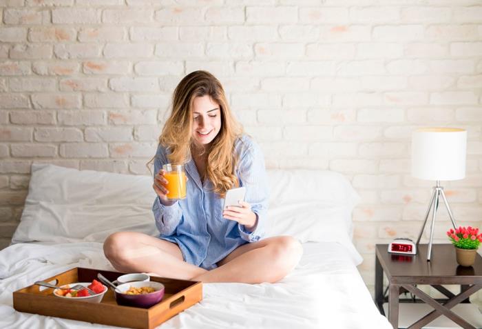 Ζώδιο ευτυχισμένο μόνο ανεξάρτητο μοναχικό νεαρό κορίτσι που κάθεται στο κρεβάτι τρώγοντας φρούτα επικοινωνεί με το κινητό τηλέφωνο