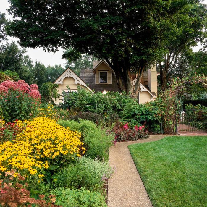 μικρό σχήμα κήπου όμορφο σπίτι πολλά πολύχρωμα λουλούδια κήπου μονοπάτι φράχτη πράσινοι θάμνοι ψηλό δέντρο